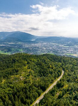 Luftaufnahme der Straße mit Wald und Bergen und Villach im Hintergrund | © villacher-alpenstrasse.at/Stabentheiner